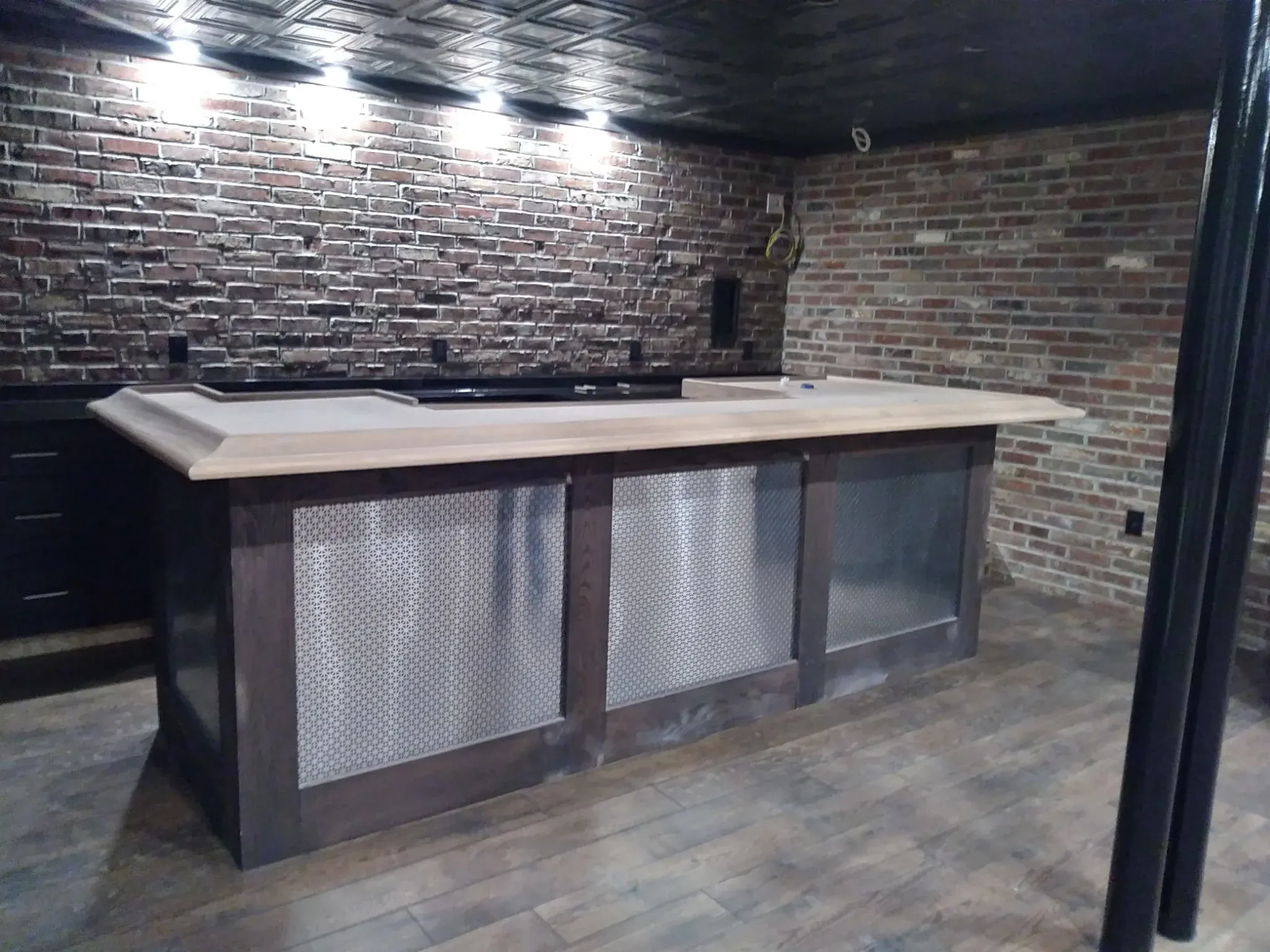 Custom basement speakeasy bar with exposed brick walls, diamond-mesh metal cabinet inserts and pressed-tin ceiling by Zavala & Sons